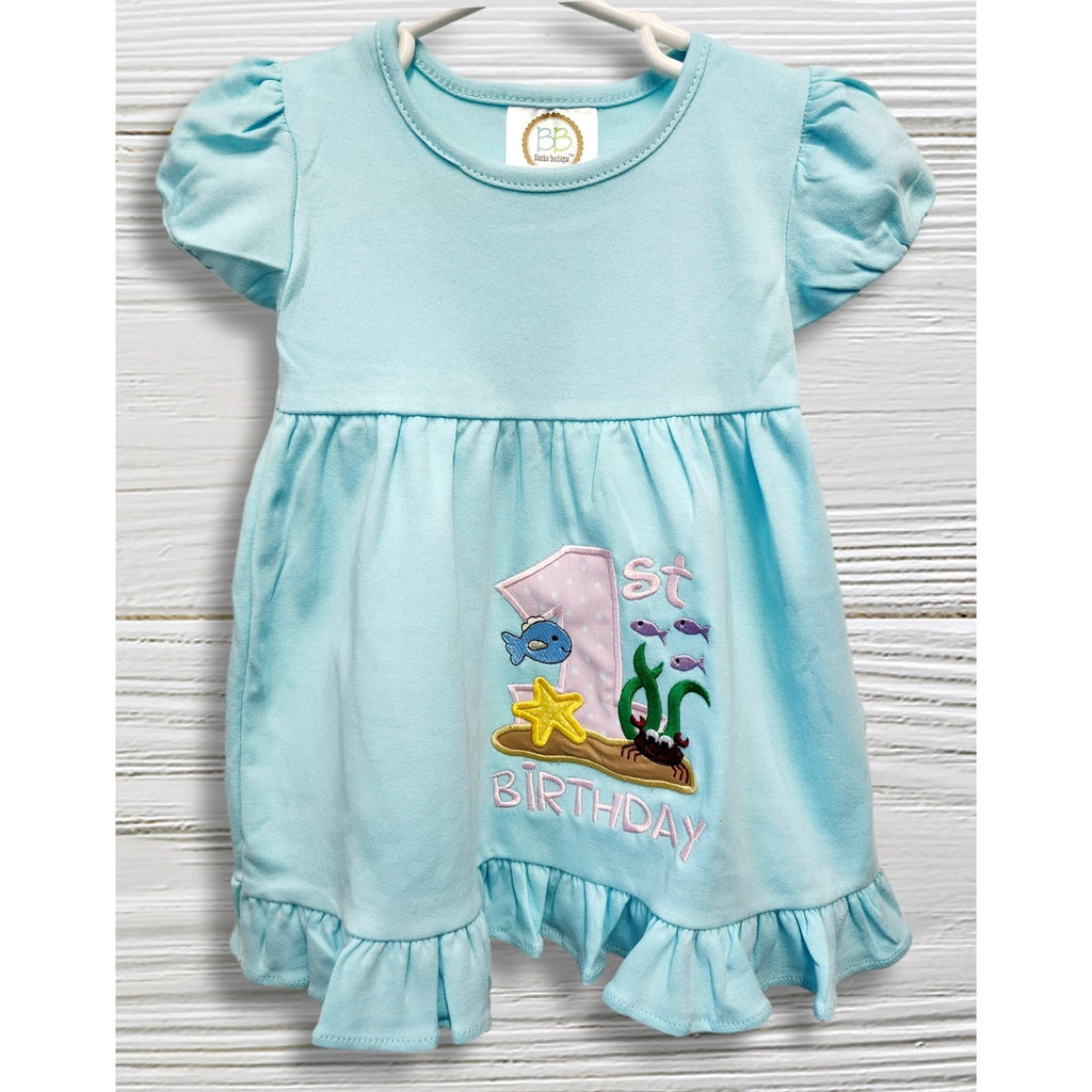Baby in a light blue dress with an '1st Birthday' design, sitting on a white rug.