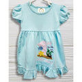 Baby in a light blue dress with an '1st Birthday' design, sitting on a white rug.