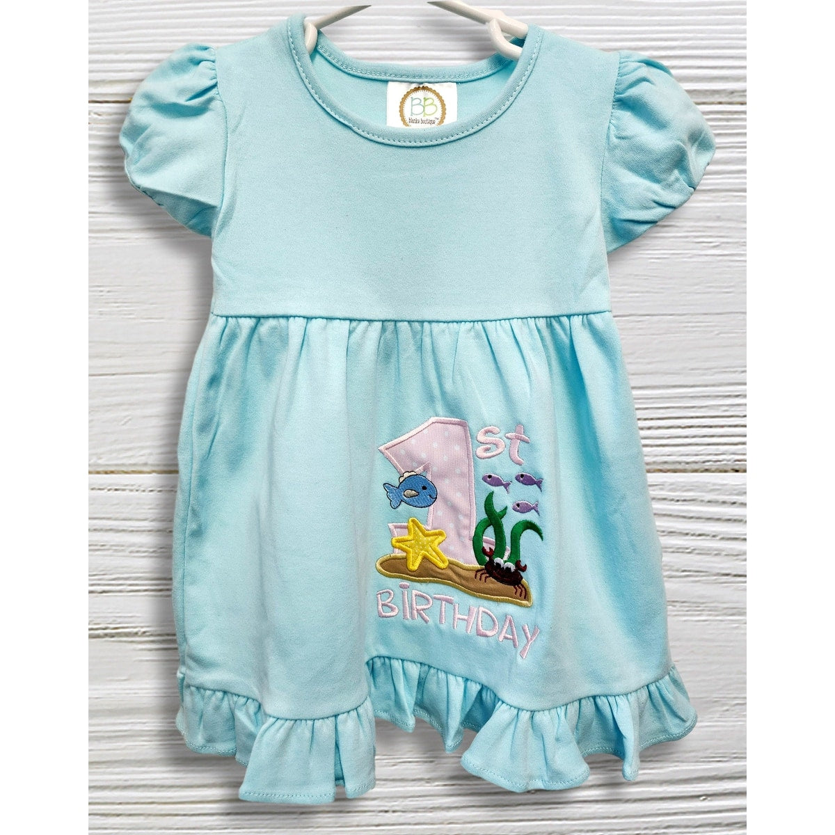 Baby in a light blue dress with an '1st Birthday' design, sitting on a white rug.