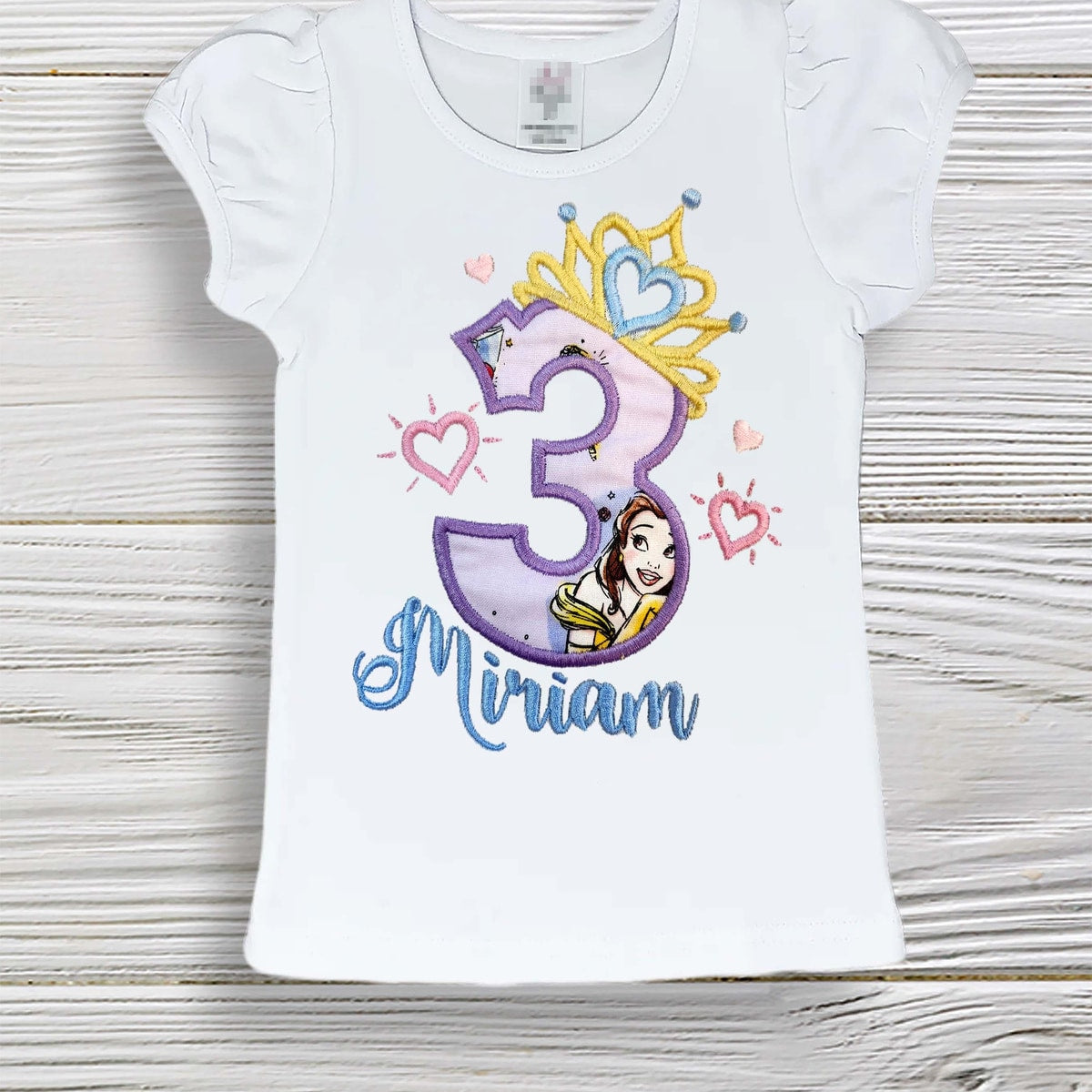 White t-shirt with a colorful number 3 design featuring a princess, on a wooden surface.