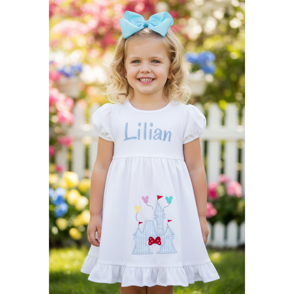 Young girl wearing a white dress with a castle design and blue bow, standing in a garden.