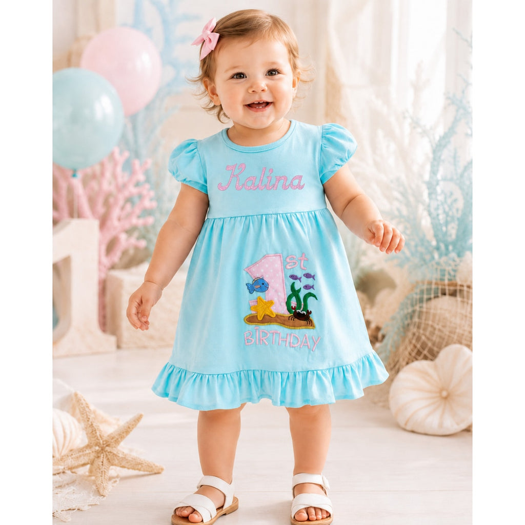 Child wearing a blue dress with '1st Birthday' and name on, standing in a decorated room.