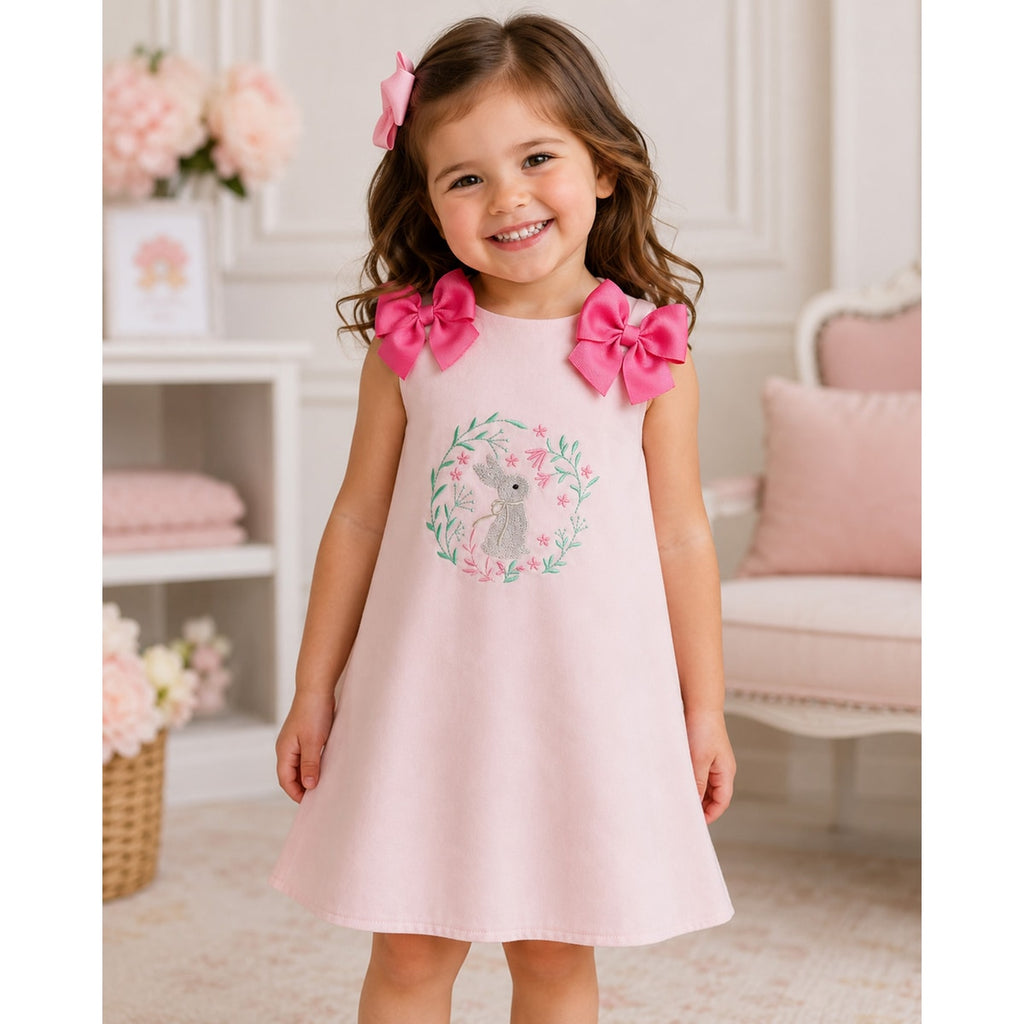 Young girl wearing a pink dress with bunny design and pink bows, standing in a softly blurred indoor setting.