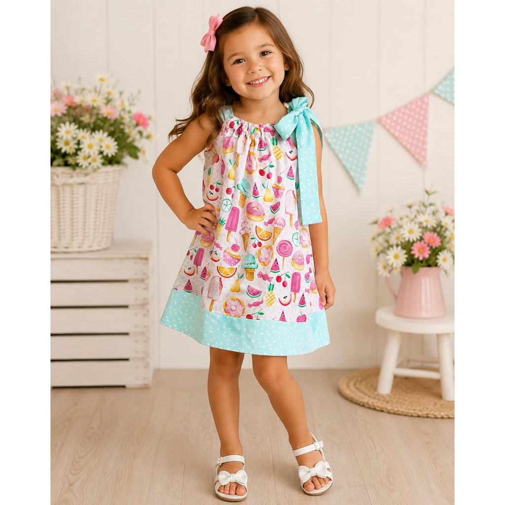 Young girl wearing a colorful dress with a pattern of ice cream cones, fruits, and other treats, standing in a bright room with flowers and a white cabinet.