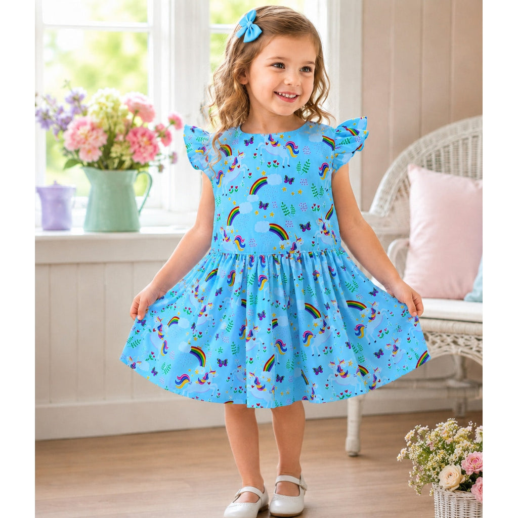 Young girl wearing a blue dress with rainbow and unicorn patterns in a bright room.
