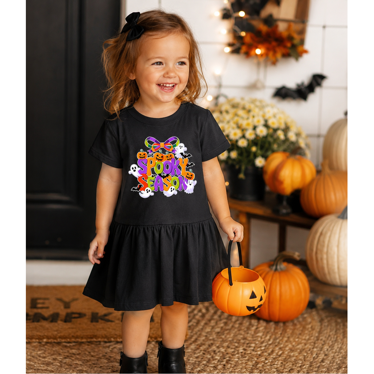 Child wearing a black Halloween-themed dress with colorful graphics, standing in front of pumpkins and flowers.