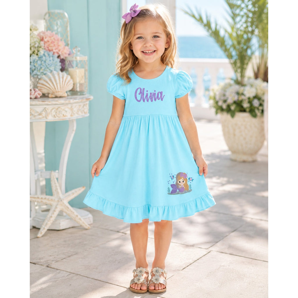 Young girl wearing a blue dress with 'Olivia' printed on it, standing on a beach.