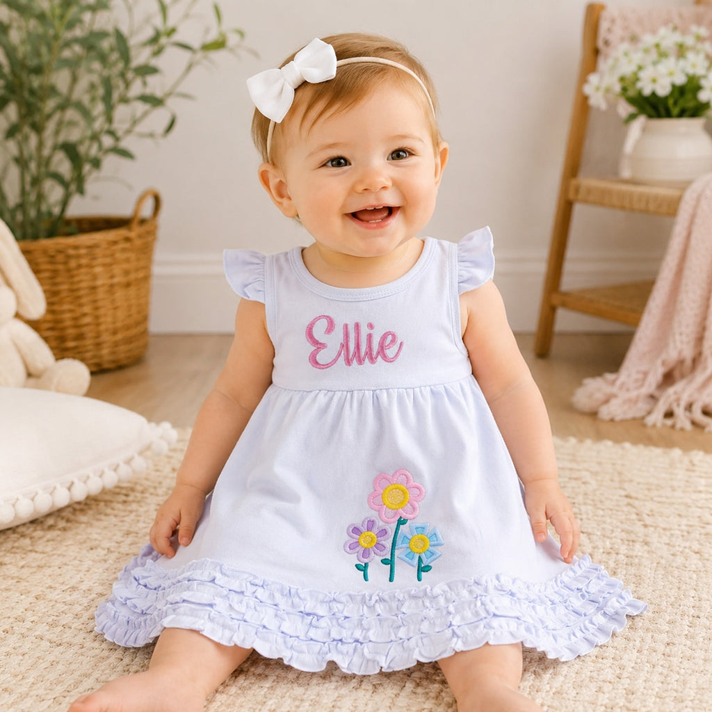 Baby girl in a white dress with floral designs and 'Ellie' printed on it, sitting on a carpeted floor.