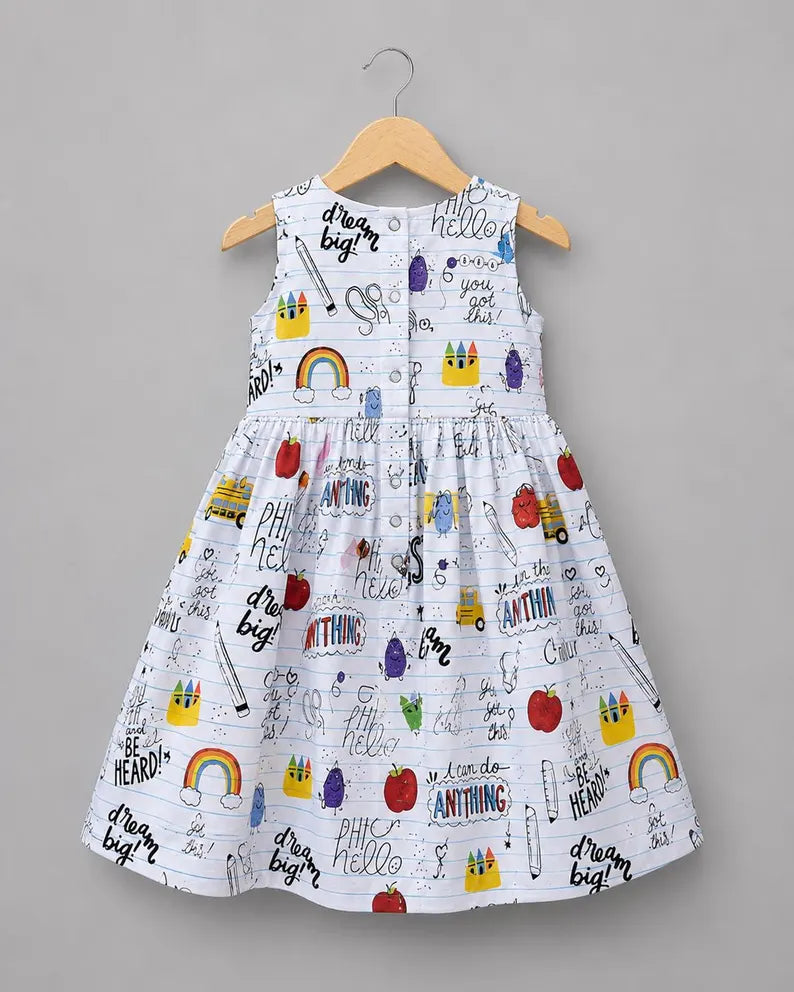 A sleeveless white children's dress with a colorful school-themed print featuring apples, rainbows, crayons, pencils, and motivational phrases like 'dream big!' and 'you can do anything,' hanging on a wooden hanger against a solid gray background.